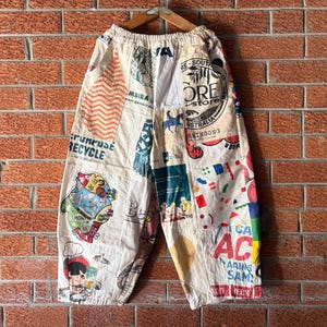 Könnte beinhalten: Cremefarbene Hose mit einem Patchwork-Design aus verschiedenen bunten Grafikdrucken, einschließlich Texten wie "RE-PURPOSE RECYCLE", "CORE STORE" und "I CAN ACT". Die Hose hat einen elastischen Bund und eine weite Beinform mit zahlreichen Illustrationen.
