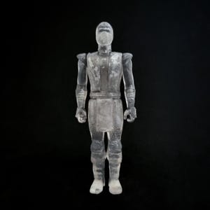 May include: A clear, translucent action figure of a ninja warrior, standing upright against a black background. The figure is detailed with a mask, armor, and boots. The toy is made of a clear material, giving it a ghostly appearance.