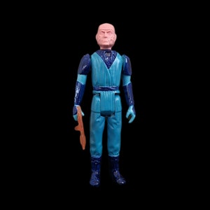 May include: Vintage action figure of a bald character in a blue and dark blue outfit, holding a brown weapon. The figure has a stern expression and is set against a black background.