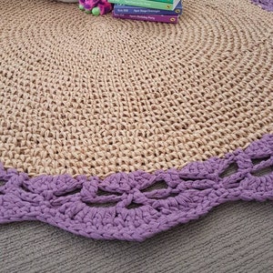 May include: A round, natural fiber rug with a purple crocheted border. The rug is on a light gray surface. There are several children's books and a stuffed animal on the rug.