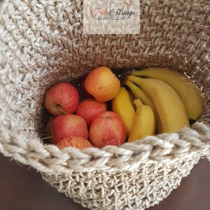 May include: A natural jute rope basket filled with red apples and yellow bananas. The basket is handmade and has a woven texture. The text "Fibs Strings" is visible on the top of the basket.