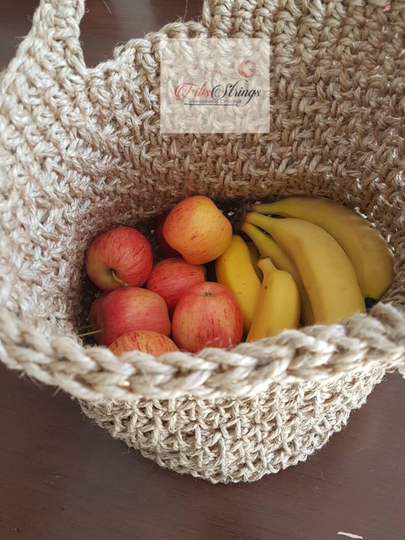 May include: A natural jute rope basket filled with red apples and yellow bananas. The basket is handmade and has a woven texture. The text "Fibs Strings" is visible on the top of the basket.