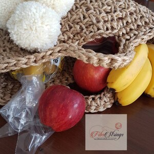 May include: A brown crocheted tote bag with white pom poms on the top. The bag is filled with fruit, including bananas and apples. The bag is sitting on a wooden surface. The text "Fibs Strings Handmade Crochet" is visible in the bottom right corner of the image.