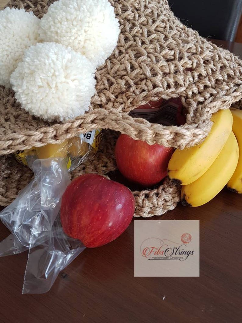 May include: A brown crocheted tote bag with white pom poms on the top. The bag is filled with fruit, including bananas and apples. The bag is sitting on a wooden surface. The text "Fibs Strings Handmade Crochet" is visible in the bottom right corner of the image.