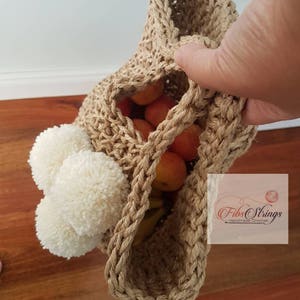 May include: A beige crocheted tote bag with white pom poms on the side. The bag is filled with fruit and is being held by a hand. The bag has the text "Fibs Strings Handmade Crochet" on the side.