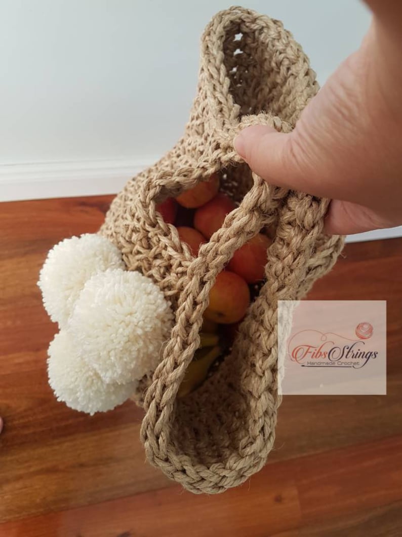 May include: A beige crocheted tote bag with white pom poms on the side. The bag is filled with fruit and is being held by a hand. The bag has the text "Fibs Strings Handmade Crochet" on the side.