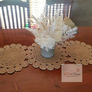 May include: Three round, crocheted, beige placemats with a decorative, scalloped edge. The placemats are arranged on a wooden table with a vase of dried white flowers in the center.