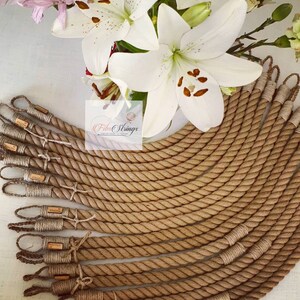 May include: A collection of beige rope handles with looped ends, each featuring a small, rectangular tag. The handles are arranged in a semi-circular pattern. White lilies and other flowers are in the background.