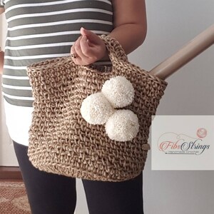 May include: A brown crocheted tote bag with three white pom-poms attached to the front. The bag has a short handle and is made of natural jute fiber. The text "Fibs Strings Handmade Crochet" is visible on the bag.