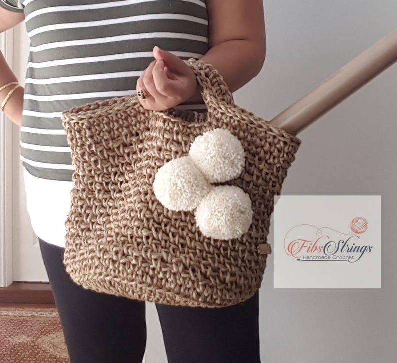 May include: A brown crocheted tote bag with three white pom-poms attached to the front. The bag has a short handle and is made of natural jute fiber. The text "Fibs Strings Handmade Crochet" is visible on the bag.