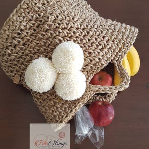 May include: A brown crocheted tote bag with white pom poms. The bag is filled with fruit, including apples and bananas. The bag is sitting on a wooden surface. The text "Fibs Strings Handmade Crochet" is visible on a white tag.