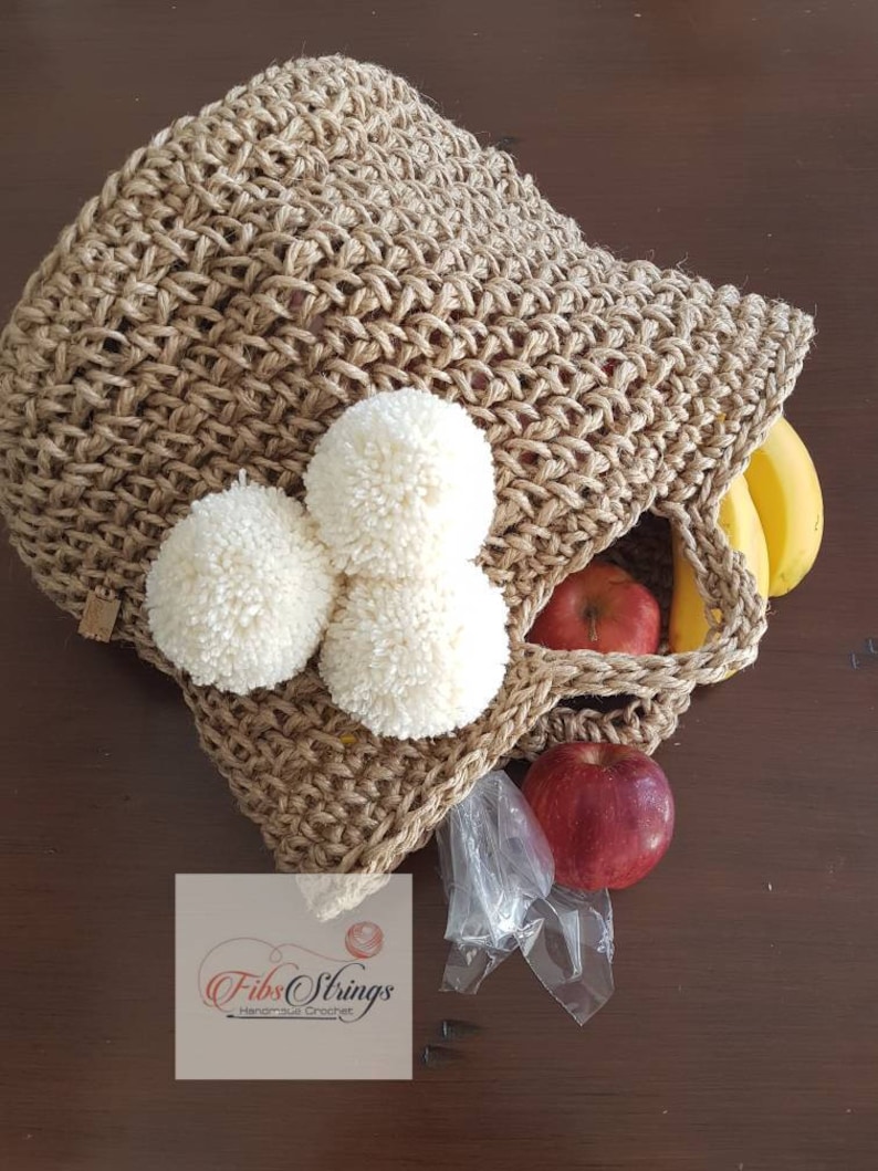 May include: A brown crocheted tote bag with white pom poms. The bag is filled with fruit, including apples and bananas. The bag is sitting on a wooden surface. The text "Fibs Strings Handmade Crochet" is visible on a white tag.