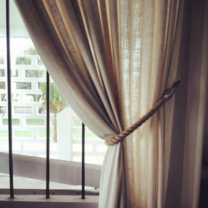 May include: Beige linen curtains tied back with a thick rope tieback. The curtains hang in front of a window with black bars and a white wall. The curtains are slightly translucent, allowing light to filter through.