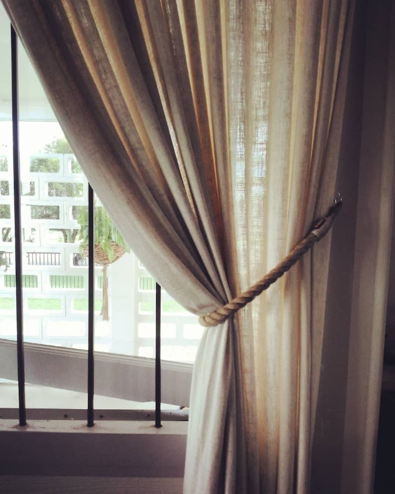 May include: Beige linen curtains tied back with a thick rope tieback. The curtains hang in front of a window with black bars and a white wall. The curtains are slightly translucent, allowing light to filter through.