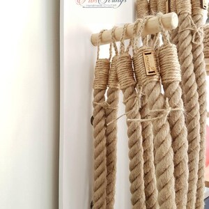 May include: Close-up of several thick, beige rope strands hanging from wooden dowels. Each rope is knotted at the top and has a small wooden tag. The ropes are part of a handmade creation, as indicated by the text "Fib Strings Handmade Crochet".