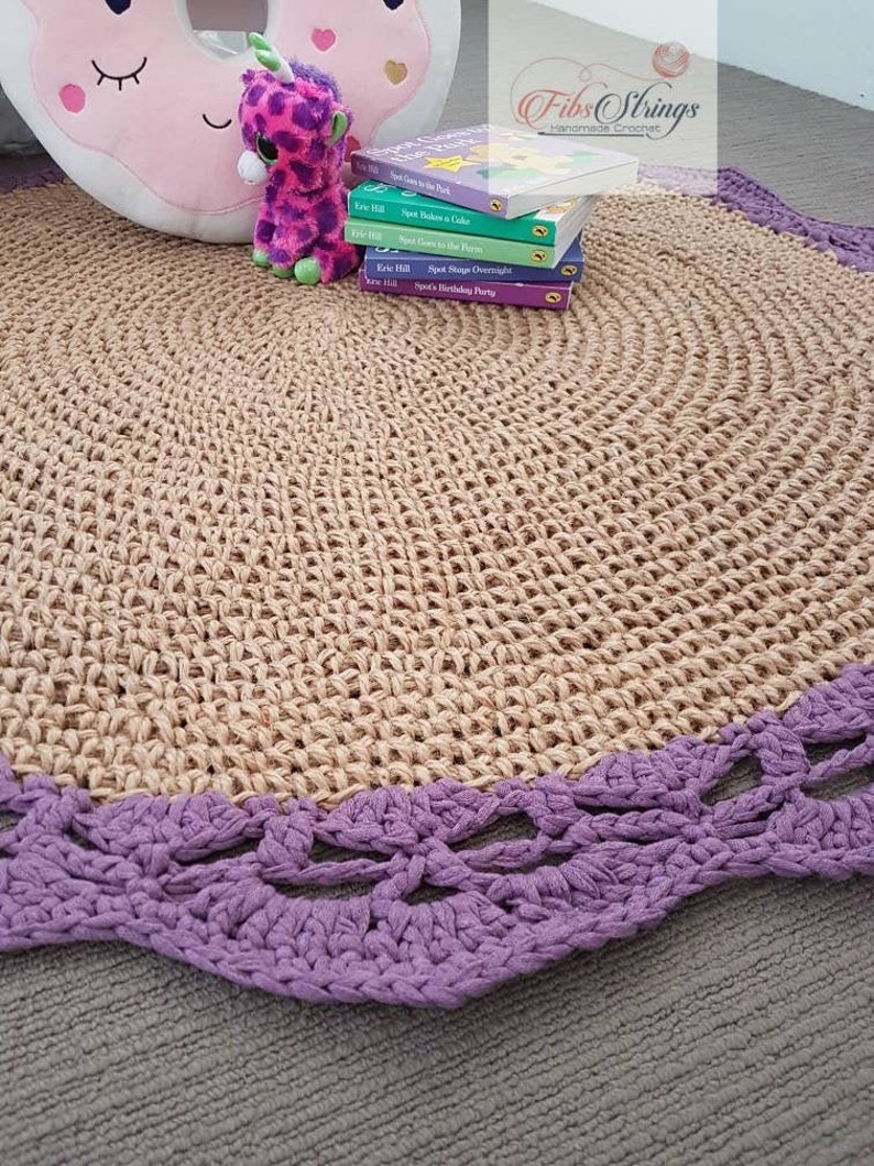 May include: A round, natural fiber rug with a purple crocheted border. The rug is on a light gray surface. There are several children's books and a stuffed animal on the rug.