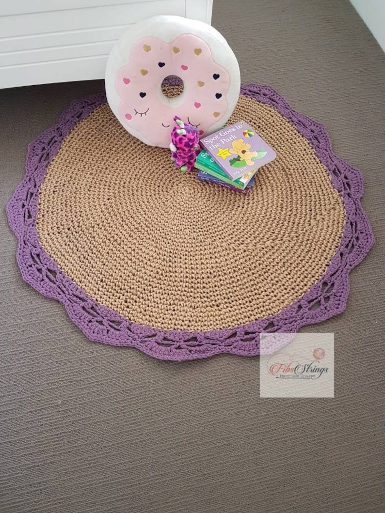 May include: A round, tan crocheted rug with a purple crocheted border. The rug is on a gray floor. A donut-shaped pillow with pink and gold accents is on the rug, along with a stack of children's books.