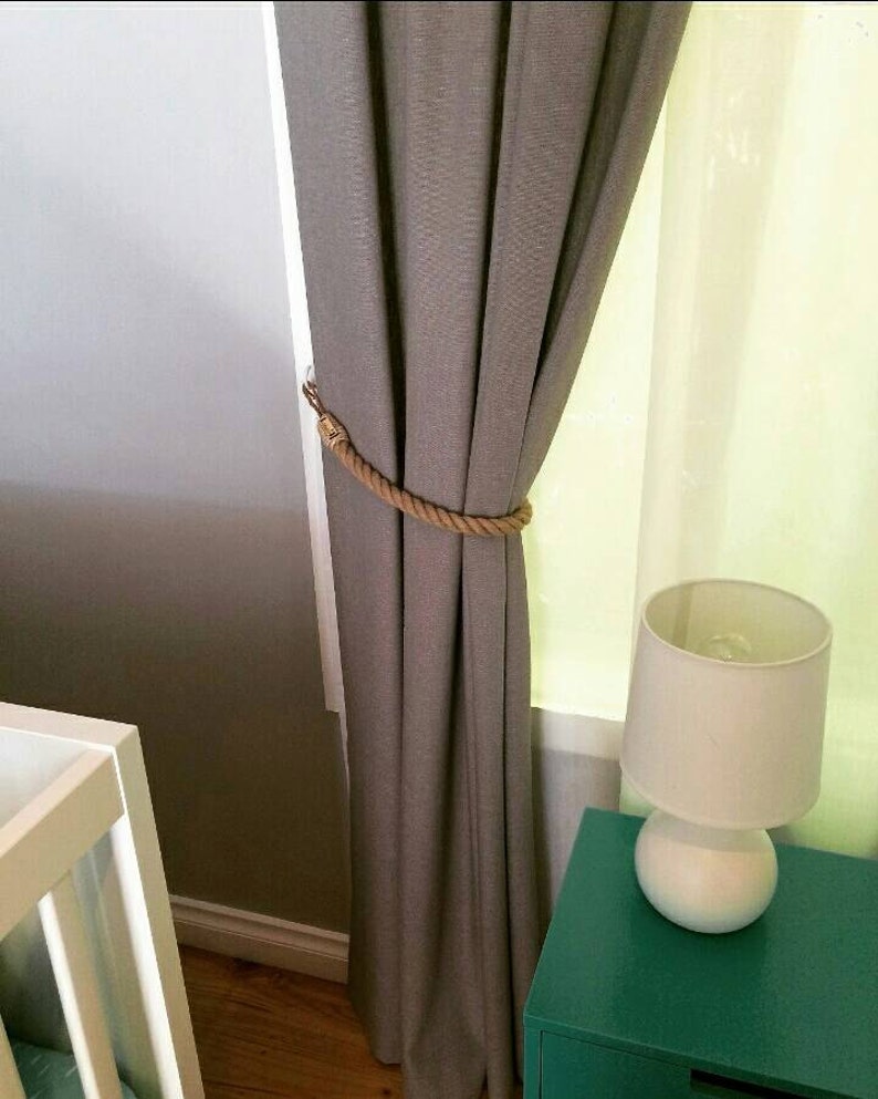 May include: A gray curtain held by a rope tieback. The curtain is next to a white wall and a green nightstand with a white lamp. A white crib is visible in the corner.