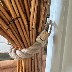 May include: A natural bamboo curtain held by a thick, woven rope tieback. The tieback is wrapped around the bamboo rods and secured with a small, rectangular tag. The bamboo rods are a light brown color.