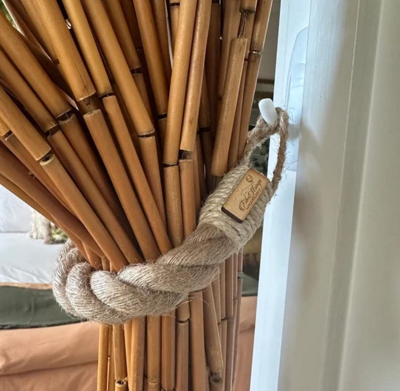 May include: A natural bamboo curtain held by a thick, woven rope tieback. The tieback is wrapped around the bamboo rods and secured with a small, rectangular tag. The bamboo rods are a light brown color.