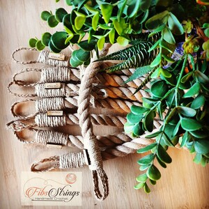 May include: Handmade crochet rope coasters with small wooden tags. The coasters are made of thick, natural-colored rope and are arranged on a wooden surface. Green foliage is in the background. The logo "Fib Strings Handmade Crochet" is visible.