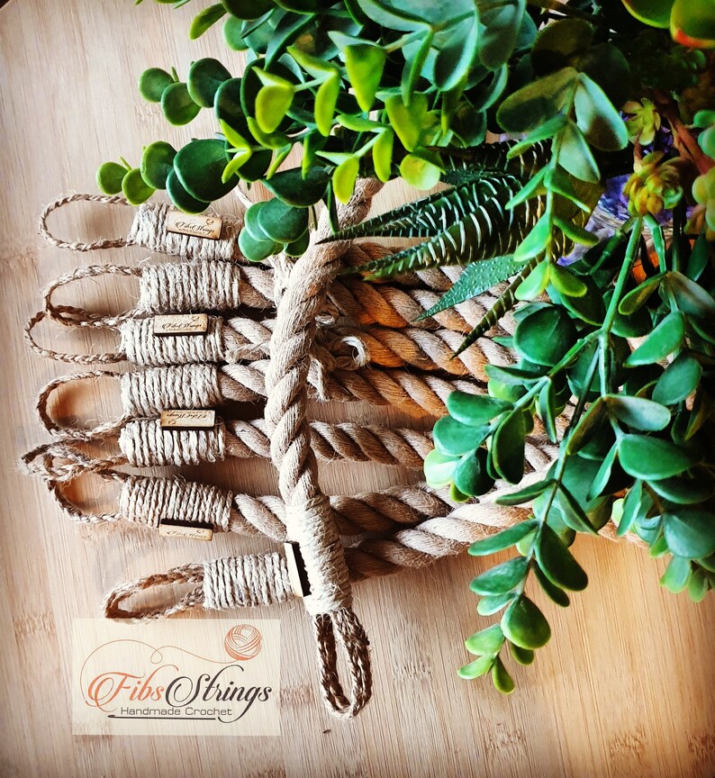 May include: Handmade crochet rope coasters with small wooden tags. The coasters are made of thick, natural-colored rope and are arranged on a wooden surface. Green foliage is in the background. The logo "Fib Strings Handmade Crochet" is visible.