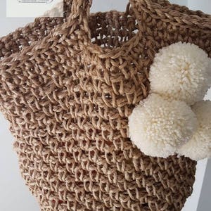 May include: A large, brown, crocheted jute tote bag with white pom poms attached to the side. The bag has a wide, sturdy handle. The text "Fibs Strings Handmade Crochet" is visible in the top left corner of the image.