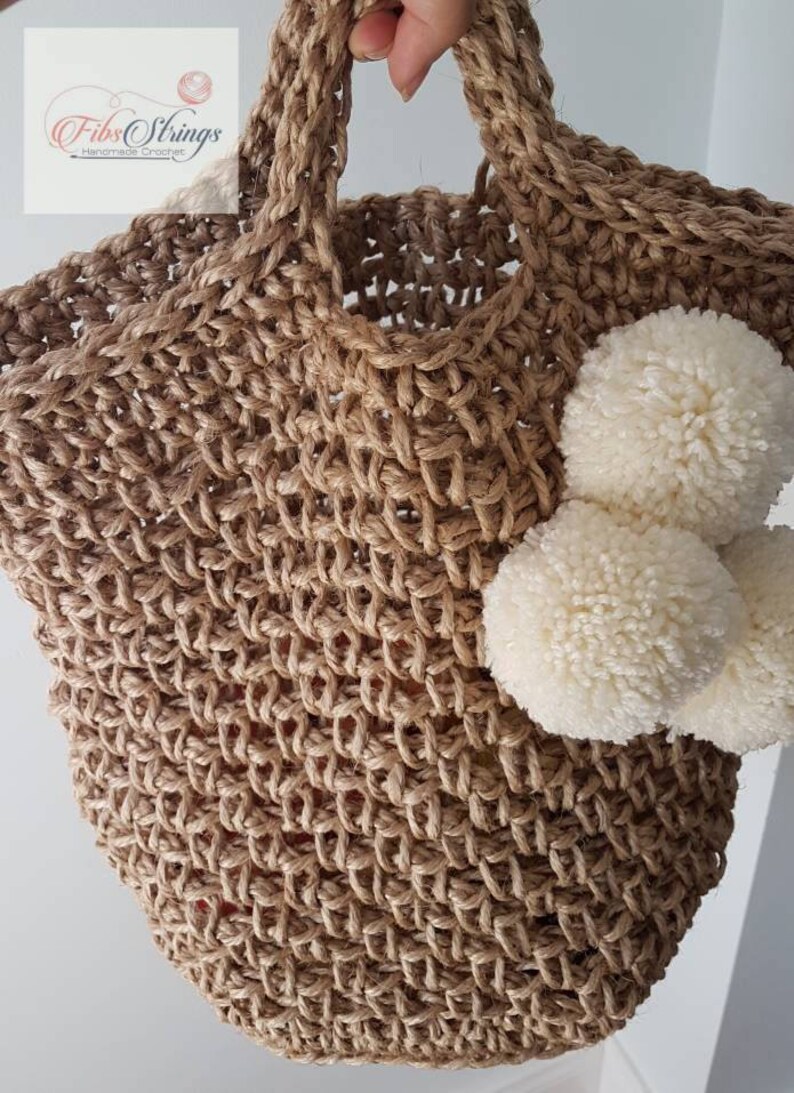 May include: A large, brown, crocheted jute tote bag with white pom poms attached to the side. The bag has a wide, sturdy handle. The text "Fibs Strings Handmade Crochet" is visible in the top left corner of the image.