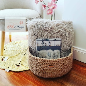 May include: A beige crochet storage basket with a fluffy gray throw blanket and a magazine inside. The basket is sitting on a yellow crochet rug in front of a white chair.