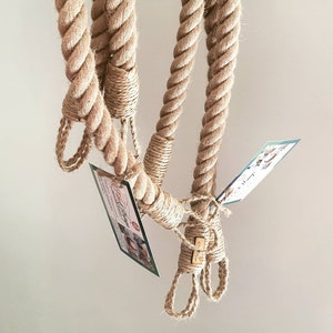 May include: Close-up of thick, natural-colored rope with loops and knots. The rope is tightly wound and appears to be made of a coarse fiber. Small tags are attached to the rope. The background is a plain, light color.