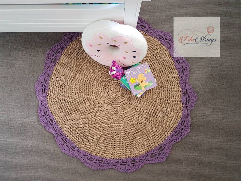 May include: A round, tan crocheted rug with a purple crocheted border. The rug has a donut-shaped pillow on top of it, and a book titled "Spot Goes to the Park" is lying on the rug.