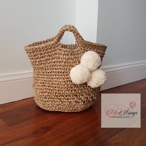 May include: A natural jute tote bag with a woven texture and white pom poms. The bag has a single handle and is handmade. The text "Fibs Strings Handmade Crochet" is visible in the lower right corner of the image.