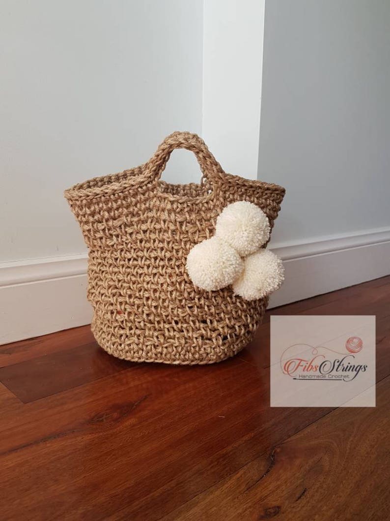 May include: A natural jute tote bag with a woven texture and white pom poms. The bag has a single handle and is handmade. The text "Fibs Strings Handmade Crochet" is visible in the lower right corner of the image.