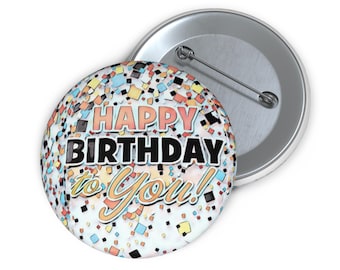 May include: A round badge with the words "HAPPY BIRTHDAY to You!" in black, orange, and yellow. The badge has a confetti design with blue, orange, and yellow squares. The badge is on a silver metal backing.