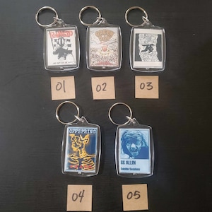May include: A collection of five clear acrylic keychains with metal rings. Each keychain features a different graphic design, including band logos and album art. The keychains are displayed on a dark surface.