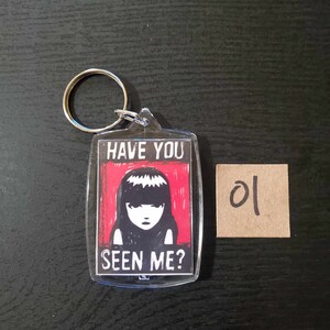 May include: Clear acrylic keychain with a silver keyring. The keychain features a graphic of a girl with black hair and the text "HAVE YOU SEEN ME?" on a red background. A small brown square with the number "01" is next to the keychain.