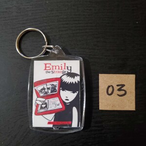 May include: A clear acrylic keychain with the text "Emily the Strange" in red. The keychain features a graphic of a girl holding a red book with illustrations. A small, square, brown tag with the number "03" is next to the keychain.