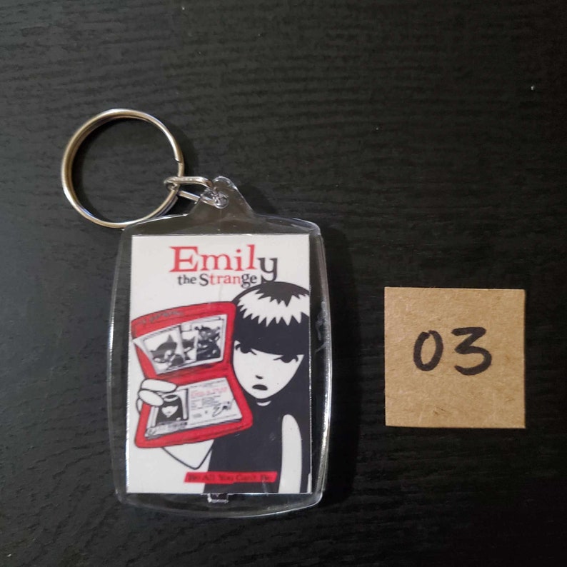 May include: A clear acrylic keychain with the text "Emily the Strange" in red. The keychain features a graphic of a girl holding a red book with illustrations. A small, square, brown tag with the number "03" is next to the keychain.