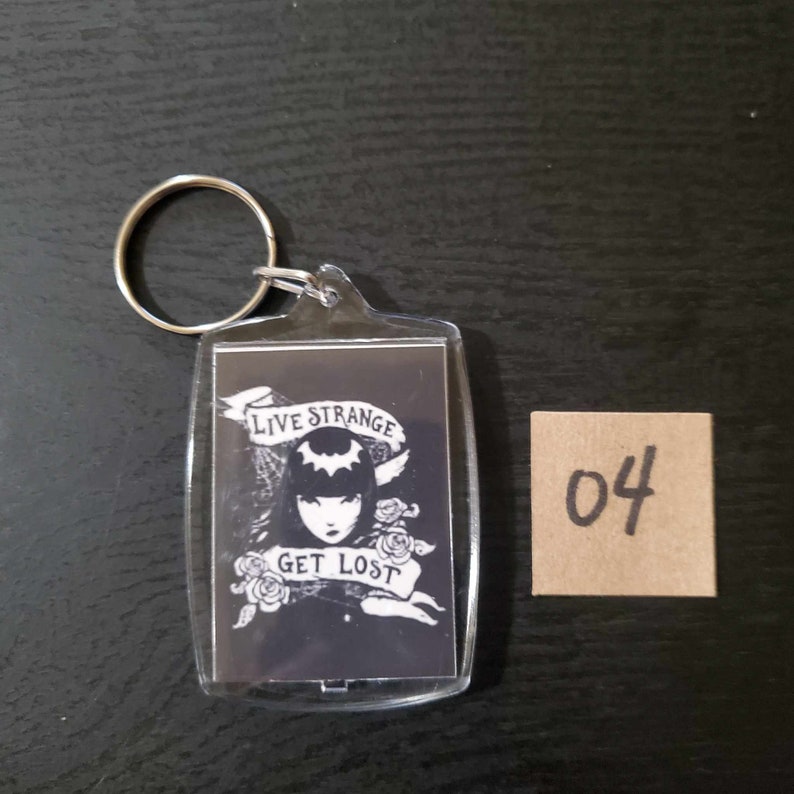 May include: A clear acrylic keyring with a silver metal ring. The keyring features a black and white graphic of a woman with the text "LIVE STRANGE GET LOST" on a banner. A small brown square with the number "04" is next to the keyring.