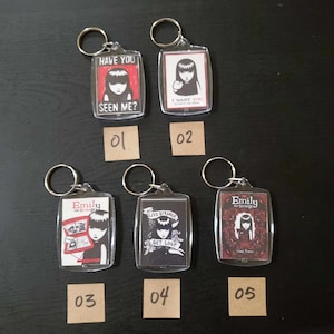 May include: A collection of five clear acrylic keychains featuring illustrations of a girl with black hair. Each keychain has a different design, including text that reads "HAVE YOU SEEN ME?", "I WANT YOU", and "LIVE STRANGE GET LOST".