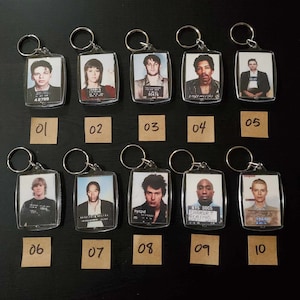May include: A collection of ten clear acrylic keychains, each featuring a mugshot-style image of a person. The keychains have silver metal rings. The images are in rectangular frames. Numbers 01 through 10 are on small brown tags below each keychain.