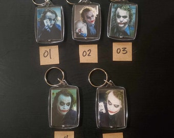 May include: Five clear acrylic keyrings featuring images of a character with white face paint and red lipstick. Each keyring has a silver metal ring. The keyrings are numbered 01, 02, 03, 04, and 05 on small brown tags.