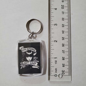 May include: A clear acrylic keyring with a rectangular image featuring a black and white illustration of a woman with the text "LIVE STRANGE GET LOST". The keyring includes a silver metal ring for attachment.