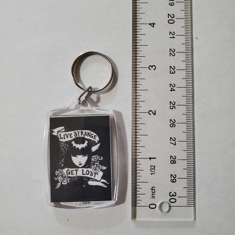 May include: A clear acrylic keyring with a rectangular image featuring a black and white illustration of a woman with the text "LIVE STRANGE GET LOST". The keyring includes a silver metal ring for attachment.