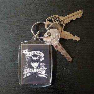 May include: A clear acrylic keyring with a black and white graphic. The graphic features a woman's face, roses, and the text "LIVE STRANGE GET LOST." Several silver keys are attached to the keyring.