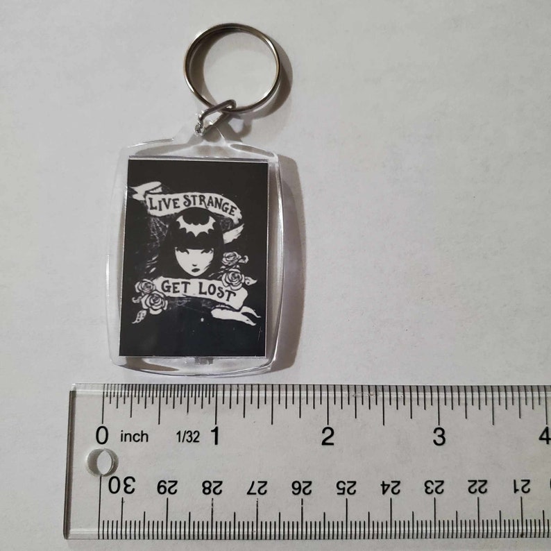 May include: A clear rectangular keyring with a silver metal ring. The keyring features a black and white graphic of a woman with the text "LIVE STRANGE GET LOST". The design includes a bat symbol and roses.
