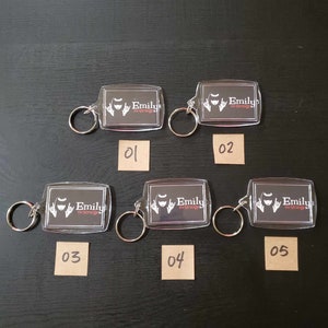 May include: Five clear acrylic keyrings with metal hoops. Each keyring features a rectangular design with a black background and the words "Emily the Strange" in white and red. The keyrings are numbered 01 to 05.