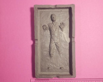 May include: A rectangular, gray sculpture depicting a person encased in a block. The figure is in a standing position with arms outstretched. The sculpture is approximately 4 inches long and is set against a pink background.