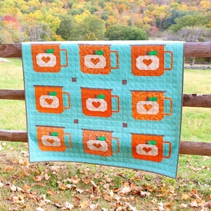 May include: A teal blue quilt with a pattern of orange mugs with hearts on them. The quilt is hanging on a wooden fence against a backdrop of fall foliage.