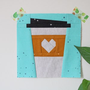 May include: A patchwork quilt block featuring a stylized coffee cup with a heart in the center. The cup is made of brown, white, and black fabric. The background is a turquoise fabric with a white star pattern.